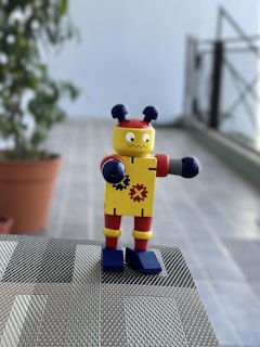 A small toy robot with colorful features stands on a woven surface. It has a yellow body decorated with gear symbols, red accents on its arms and legs, and blue shoes. Two antenna-like protrusions are on its head. The background includes a potted plant and a blurred view of a tiled floor and railing.