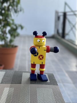 A small toy robot with colorful features stands on a woven surface. It has a yellow body decorated with gear symbols, red accents on its arms and legs, and blue shoes. Two antenna-like protrusions are on its head. The background includes a potted plant and a blurred view of a tiled floor and railing.