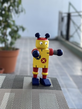 A small toy robot with colorful features stands on a woven surface. It has a yellow body decorated with gear symbols, red accents on its arms and legs, and blue shoes. Two antenna-like protrusions are on its head. The background includes a potted plant and a blurred view of a tiled floor and railing.
