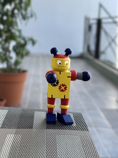 A small toy robot with colorful features stands on a woven surface. It has a yellow body decorated with gear symbols, red accents on its arms and legs, and blue shoes. Two antenna-like protrusions are on its head. The background includes a potted plant and a blurred view of a tiled floor and railing.