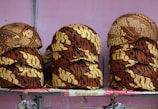 Stacks of intricately patterned caps are arranged on a shelf. The caps feature a traditional batik design with rich brown and beige colors and various motifs.