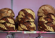 Stacks of intricately patterned caps are arranged on a shelf. The caps feature a traditional batik design with rich brown and beige colors and various motifs.