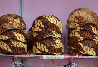 Stacks of intricately patterned caps are arranged on a shelf. The caps feature a traditional batik design with rich brown and beige colors and various motifs.