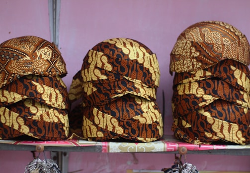 Stacks of intricately patterned caps are arranged on a shelf. The caps feature a traditional batik design with rich brown and beige colors and various motifs.