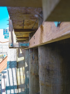 A wooden structure featuring beams and planks held together by metal brackets. Sunlight casts shadows and highlights textures on the wood. A sign and a portion of the sky are visible in the background.