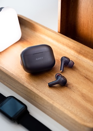 A pair of wireless earbuds and a charging case are placed on a wooden tray. Next to the tray is a smartwatch with a black band and a bright white lamp casting light onto the scene. The earbuds and case have a sleek black design with subtle branding.