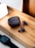 A pair of wireless earbuds and a charging case are placed on a wooden tray. Next to the tray is a smartwatch with a black band and a bright white lamp casting light onto the scene. The earbuds and case have a sleek black design with subtle branding.