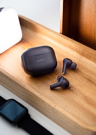 A pair of wireless earbuds and a charging case are placed on a wooden tray. Next to the tray is a smartwatch with a black band and a bright white lamp casting light onto the scene. The earbuds and case have a sleek black design with subtle branding.