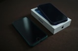 A smartphone lies on top of its box on a dark surface, with another phone placed nearby. The box displays a logo, suggesting a brand. Both phones appear to have dark screens.