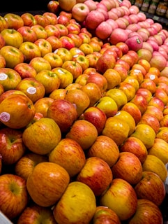 A display of colorful, shiny apples carefully arranged and stacked in a grocery store or market setting. The apples vary in shades, predominantly red and yellow, creating a vibrant and fresh appearance. Some of the apples have stickers with branding or pricing information.