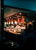 A sushi restaurant interior is captured with chefs in white uniforms preparing food behind a wooden counter. The ambience is warm with overhead pendant lighting and the presence of various kitchen items and ingredients on the counter. The restaurant has a cozy and traditional decor with wooden elements.