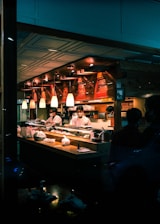 A sushi restaurant interior is captured with chefs in white uniforms preparing food behind a wooden counter. The ambience is warm with overhead pendant lighting and the presence of various kitchen items and ingredients on the counter. The restaurant has a cozy and traditional decor with wooden elements.