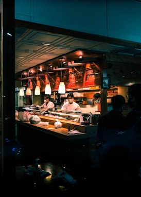 A sushi restaurant interior is captured with chefs in white uniforms preparing food behind a wooden counter. The ambience is warm with overhead pendant lighting and the presence of various kitchen items and ingredients on the counter. The restaurant has a cozy and traditional decor with wooden elements.