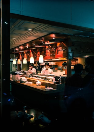 A sushi restaurant interior is captured with chefs in white uniforms preparing food behind a wooden counter. The ambience is warm with overhead pendant lighting and the presence of various kitchen items and ingredients on the counter. The restaurant has a cozy and traditional decor with wooden elements.