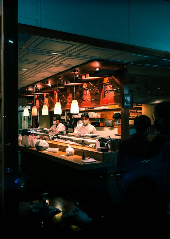 A sushi restaurant interior is captured with chefs in white uniforms preparing food behind a wooden counter. The ambience is warm with overhead pendant lighting and the presence of various kitchen items and ingredients on the counter. The restaurant has a cozy and traditional decor with wooden elements.