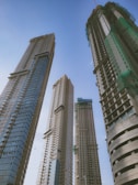 Four tall skyscrapers under construction rise against a clear blue sky. The buildings feature modern architectural designs with glass and concrete elements. Green safety nets are visible on some sections, indicating ongoing construction work.