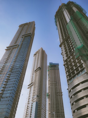 Four tall skyscrapers under construction rise against a clear blue sky. The buildings feature modern architectural designs with glass and concrete elements. Green safety nets are visible on some sections, indicating ongoing construction work.