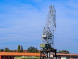 A large industrial crane stands against a clear blue sky. The crane has a metallic structure with a long arm extending upwards, equipped with a hook dangling from its end. Below the crane, there is a red-roofed building and some trees surrounding the area.