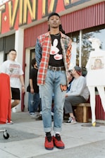 A person stands confidently in front of a vintage clothing store, wearing a plaid shirt over a graphic tee, jeans, and red sneakers. Several other individuals are in the background, some interacting and others casually pose. The storefront displays vibrant signage and mannequins wearing vintage-style outfits.
