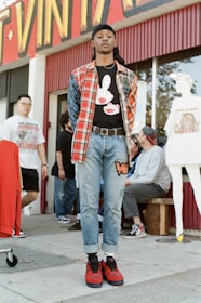 A person stands confidently in front of a vintage clothing store, wearing a plaid shirt over a graphic tee, jeans, and red sneakers. Several other individuals are in the background, some interacting and others casually pose. The storefront displays vibrant signage and mannequins wearing vintage-style outfits.