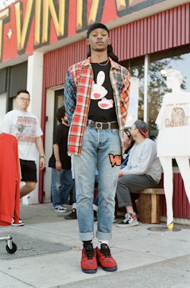 A person stands confidently in front of a vintage clothing store, wearing a plaid shirt over a graphic tee, jeans, and red sneakers. Several other individuals are in the background, some interacting and others casually pose. The storefront displays vibrant signage and mannequins wearing vintage-style outfits.