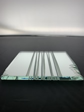 A clear, rectangular glass panel with beveled edges is lying flat on a dark surface. The panel's surface has several vertical grooves that create a pattern. Light reflects subtly off the glass, adding a hint of blue and green hues.