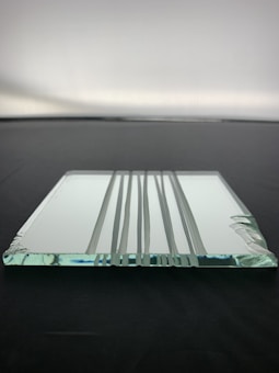 A clear, rectangular glass panel with beveled edges is lying flat on a dark surface. The panel's surface has several vertical grooves that create a pattern. Light reflects subtly off the glass, adding a hint of blue and green hues.