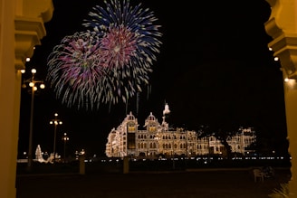 A grand, historical palace is brightly illuminated against the night sky, with elaborate fireworks burst overhead creating a colorful visual spectacle. The ornate architecture of the palace is complemented by the warm glow of countless lights outlining its structure, enhancing its majestic appearance. The scene is framed by large archways, adding depth and a sense of grandeur.