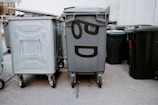 A group of large garbage bins are arranged on a street next to a building. One of the bins is decorated with painted eyes and a mouth, giving it a cartoon-like expression. The concrete ground and building wall provide a neutral urban backdrop.