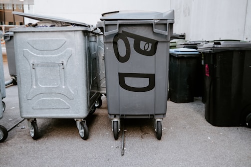 A group of large garbage bins are arranged on a street next to a building. One of the bins is decorated with painted eyes and a mouth, giving it a cartoon-like expression. The concrete ground and building wall provide a neutral urban backdrop.