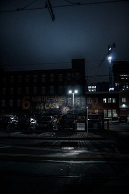 A dimly lit urban scene at night featuring a brick building with a faded sign that reads '6 MONTHS SAME AS CASH.' The street is wet, possibly from rain, and illuminated by the glow of streetlights. A construction crane is visible in the background, along with parked cars and a fenced parking area.