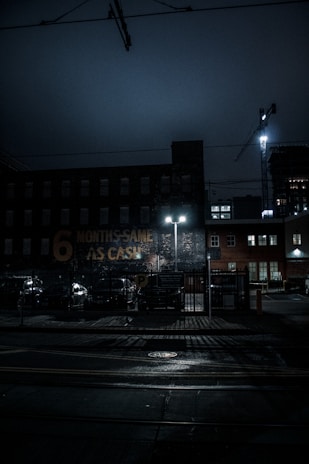 A dimly lit urban scene at night featuring a brick building with a faded sign that reads '6 MONTHS SAME AS CASH.' The street is wet, possibly from rain, and illuminated by the glow of streetlights. A construction crane is visible in the background, along with parked cars and a fenced parking area.