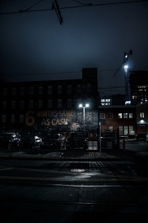 A dimly lit urban scene at night featuring a brick building with a faded sign that reads '6 MONTHS SAME AS CASH.' The street is wet, possibly from rain, and illuminated by the glow of streetlights. A construction crane is visible in the background, along with parked cars and a fenced parking area.