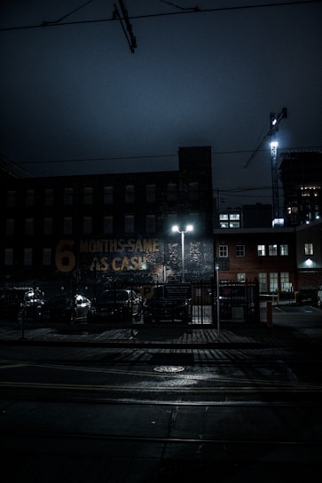 A dimly lit urban scene at night featuring a brick building with a faded sign that reads '6 MONTHS SAME AS CASH.' The street is wet, possibly from rain, and illuminated by the glow of streetlights. A construction crane is visible in the background, along with parked cars and a fenced parking area.