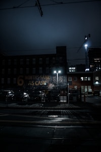 A dimly lit urban scene at night featuring a brick building with a faded sign that reads '6 MONTHS SAME AS CASH.' The street is wet, possibly from rain, and illuminated by the glow of streetlights. A construction crane is visible in the background, along with parked cars and a fenced parking area.