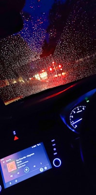 A car interior viewed from the driver's seat during nighttime. Raindrops are scattered across the windshield, creating a distorted view of red tail lights from vehicles ahead. The dashboard displays Spotify playing a song.