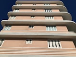 A multi-story building with a distinctive art deco architectural style features smooth, curved edges and a pastel pink facade. The structure has multiple white-framed windows evenly distributed across its levels, against a clear blue sky.