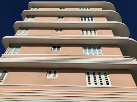 A multi-story building with a distinctive art deco architectural style features smooth, curved edges and a pastel pink facade. The structure has multiple white-framed windows evenly distributed across its levels, against a clear blue sky.