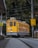 A yellow and blue tram moves along the track surrounded by lush green trees and various overhead electrical wires. There are a few signs around, one of which instructs to reduce speed. The tram is number 23 and has an indication that it goes to Dois Irmãos.
