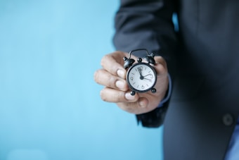 A person in a suit holding a small, classic black alarm clock with white clock face. The background is a solid light blue color, creating a contrast with the dark suit and clock.