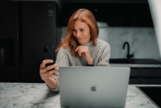 A woman with auburn hair is sitting at a marble countertop, holding a smartphone and looking at it while smiling. She has a light gray knitted sweater on and is seated in front of an open silver laptop, possibly in a kitchen setting with dark cabinetry and a sleek modern design.