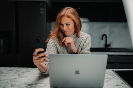 A woman with auburn hair is sitting at a marble countertop, holding a smartphone and looking at it while smiling. She has a light gray knitted sweater on and is seated in front of an open silver laptop, possibly in a kitchen setting with dark cabinetry and a sleek modern design.