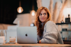 A person with long brown hair wearing a cozy, oversized gray sweater sits at a wooden table in a warmly lit cafe. In front of them is an open laptop, and to the side, a glass of latte and a small plate with a pastry. The background features blurred decorative elements and soft lighting, creating a relaxed atmosphere.