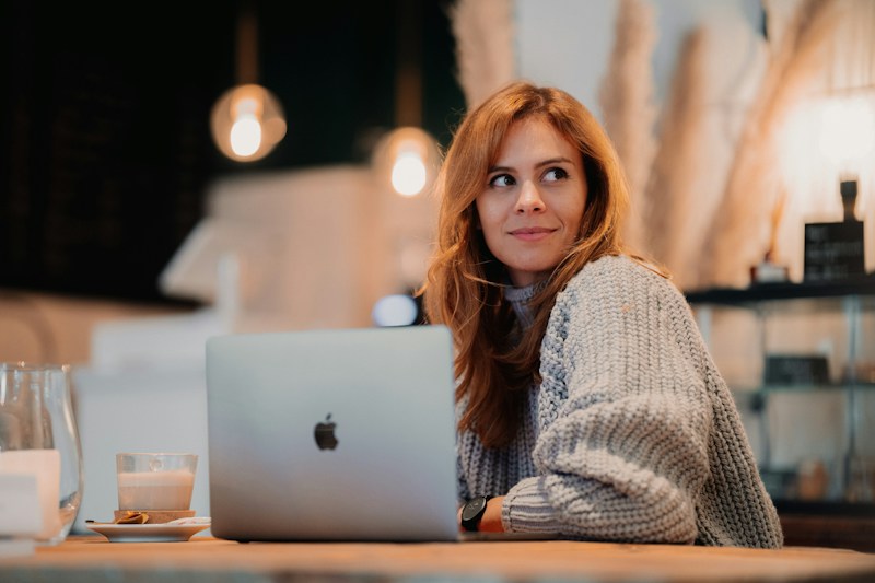 A person with long brown hair wearing a cozy, oversized gray sweater sits at a wooden table in a warmly lit cafe. In front of them is an open laptop, and to the side, a glass of latte and a small plate with a pastry. The background features blurred decorative elements and soft lighting, creating a relaxed atmosphere.