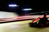 A go-kart speeding along a track at night, with blurred motion lines emphasizing the sense of rapid movement. Bright streaks of red and white lights create a dynamic and energetic atmosphere.