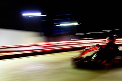A go-kart speeding along a track at night, with blurred motion lines emphasizing the sense of rapid movement. Bright streaks of red and white lights create a dynamic and energetic atmosphere.