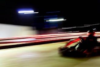 A go-kart speeding along a track at night, with blurred motion lines emphasizing the sense of rapid movement. Bright streaks of red and white lights create a dynamic and energetic atmosphere.