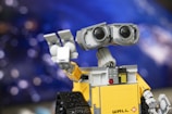 A LEGO model depicts a robot with large binocular-like eyes and a square, yellow body. The robot's arm is raised as if waving or holding something. The background features a blurry, bokeh effect with various shades of blue and purple resembling a starry sky or space.