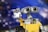 A LEGO model depicts a robot with large binocular-like eyes and a square, yellow body. The robot's arm is raised as if waving or holding something. The background features a blurry, bokeh effect with various shades of blue and purple resembling a starry sky or space.