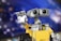 A LEGO model depicts a robot with large binocular-like eyes and a square, yellow body. The robot's arm is raised as if waving or holding something. The background features a blurry, bokeh effect with various shades of blue and purple resembling a starry sky or space.